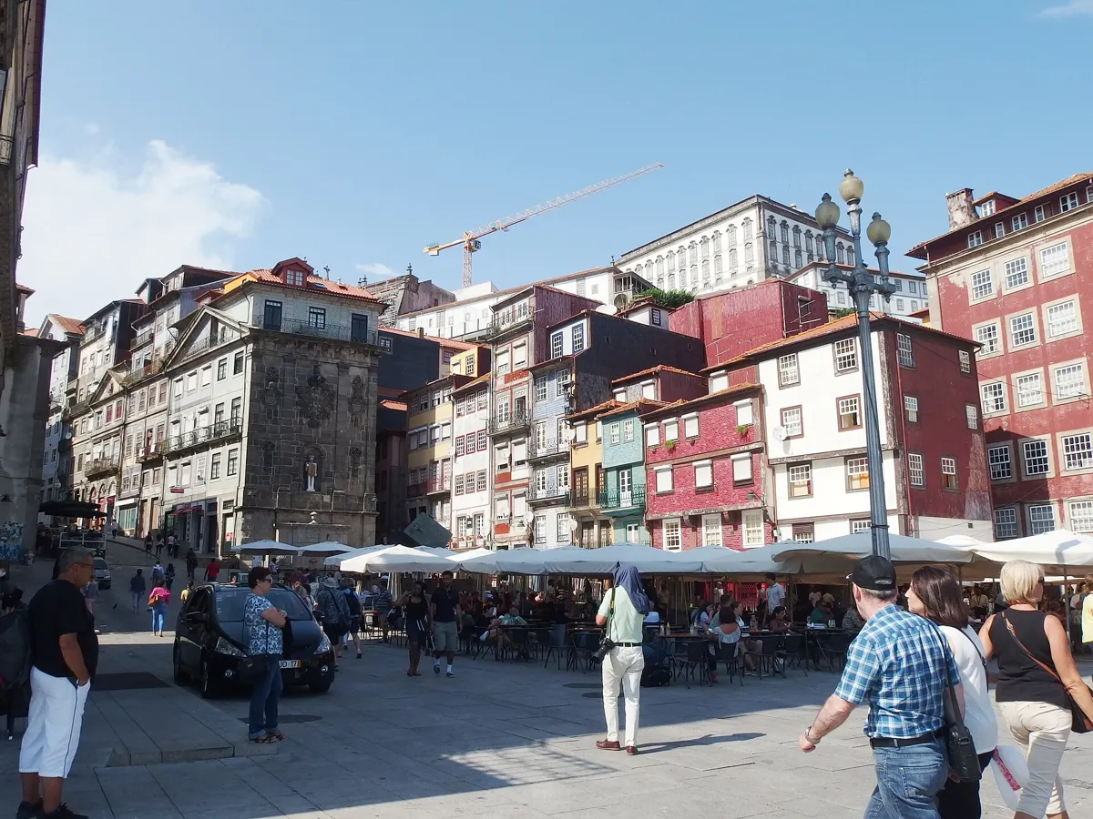 A busy riverside square with café umbrellas, a street lamp and tall narrow houses painted in red, yellow and white