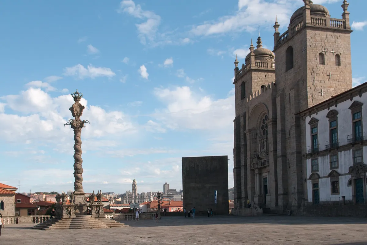 A wide paved terrace with a twisted stone column on a stepped base, and the cathedral's two square towers on the right under a blue sky