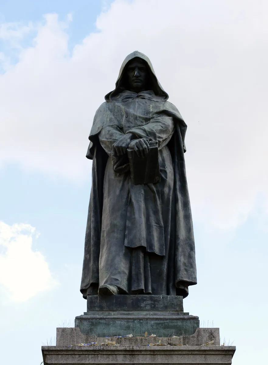 The dark bronze statue of Giordano Bruno in a monk's hooded habit, hands folded over a book, on a stone pedestal against a pale sky