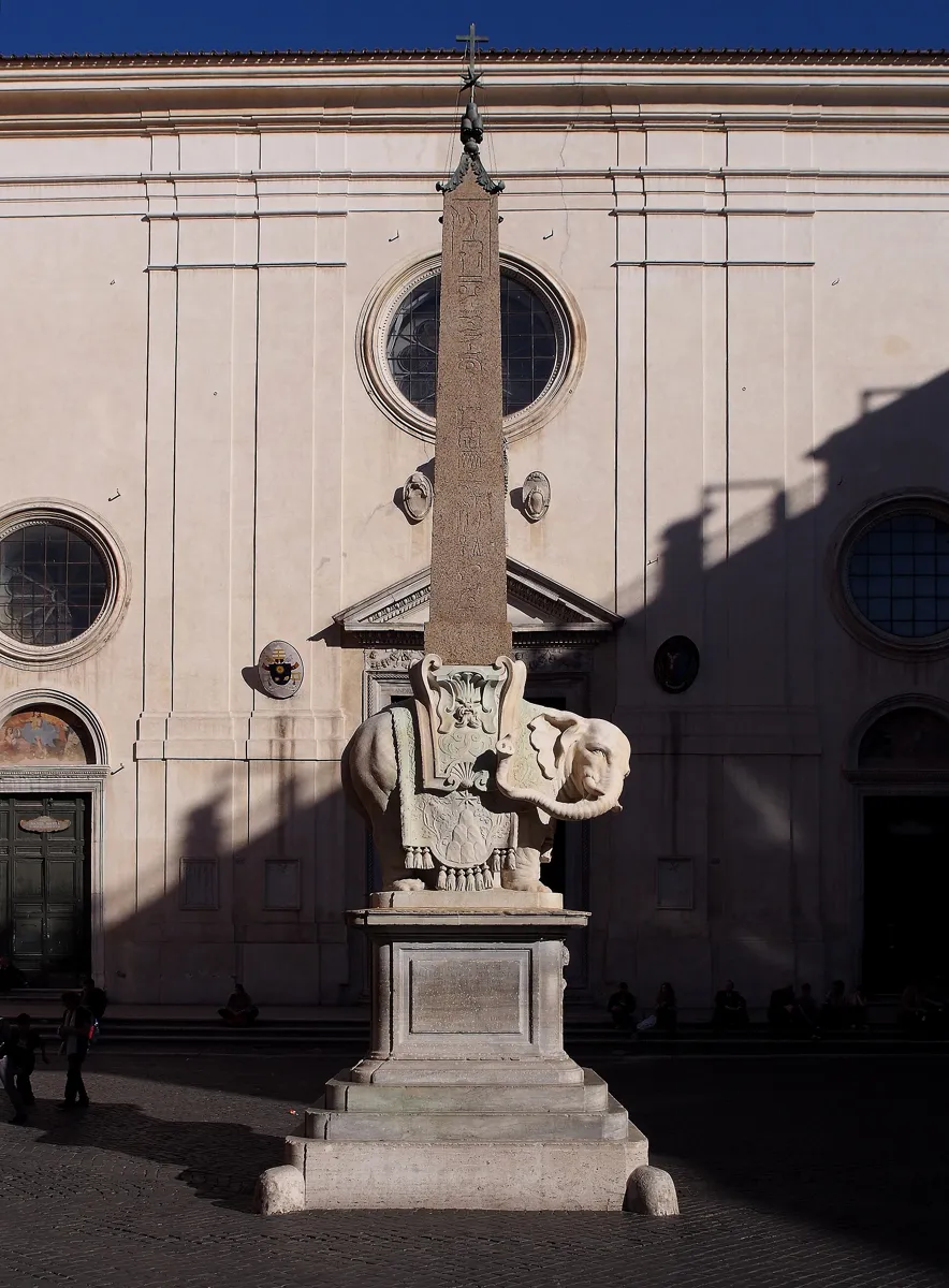 A small white marble elephant with a decorated saddle cloth carries a tall, thin Egyptian obelisk on its back, in front of the plain pale front of Santa Maria sopra Minerva