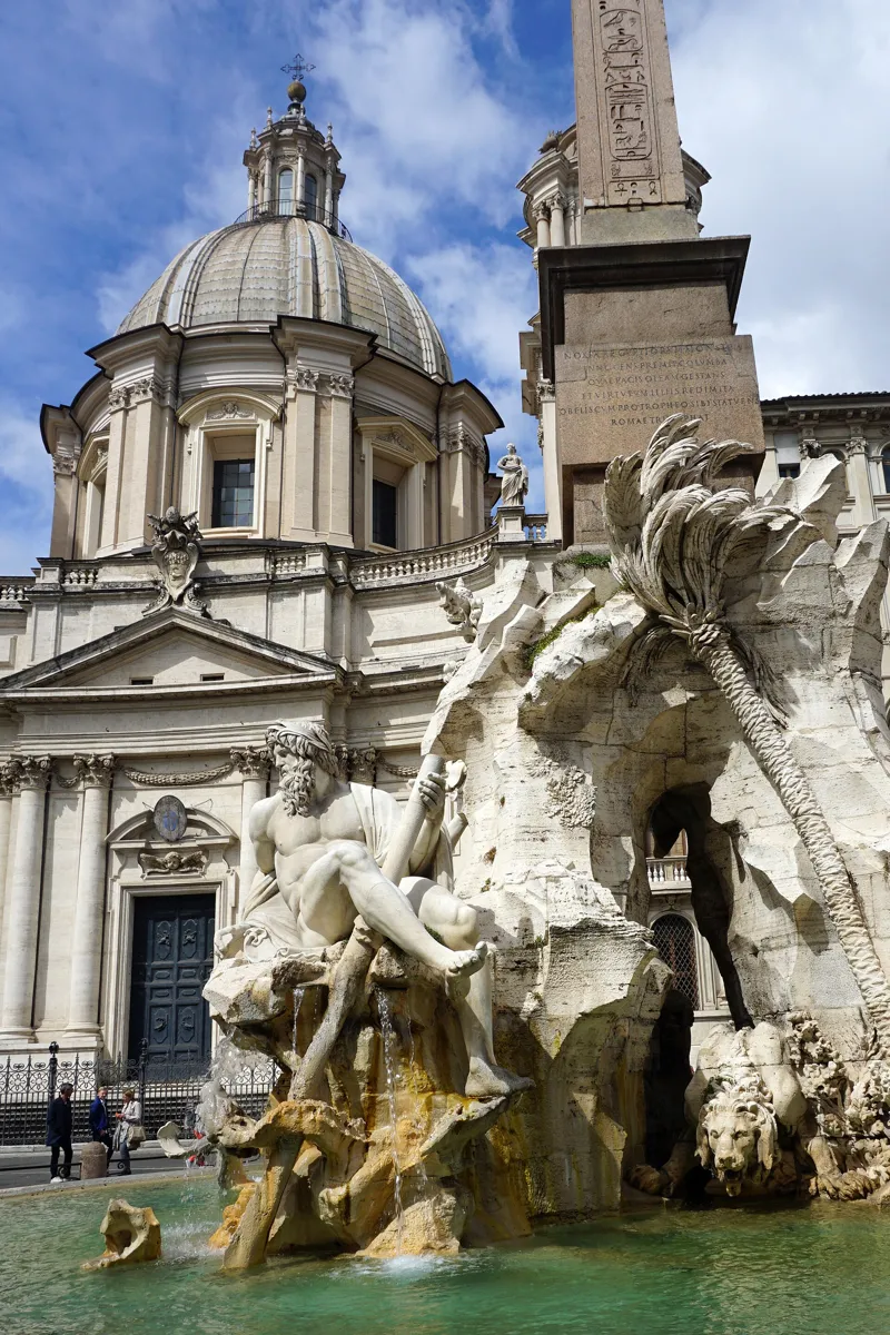 A marble river god holding a long oar sits on the rocks of Bernini's fountain, with a carved palm tree, a lion and the obelisk beside him and the dome of Sant'Agnese in Agone behind
