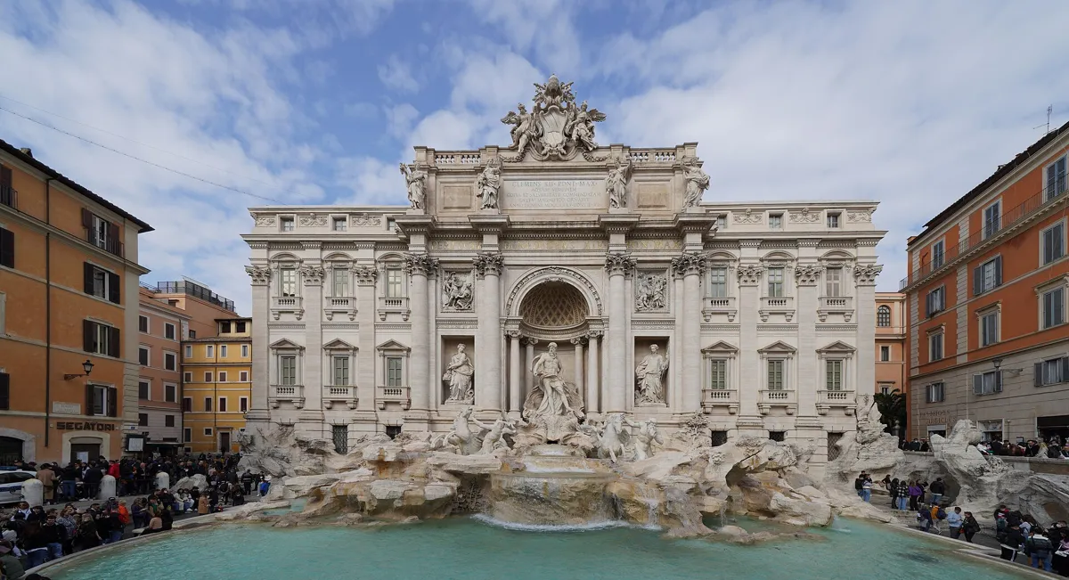 The Trevi Fountain seen from across its turquoise basin: a white palace front with a central arch and the statue of Oceanus above carved rocks, with crowds on both sides